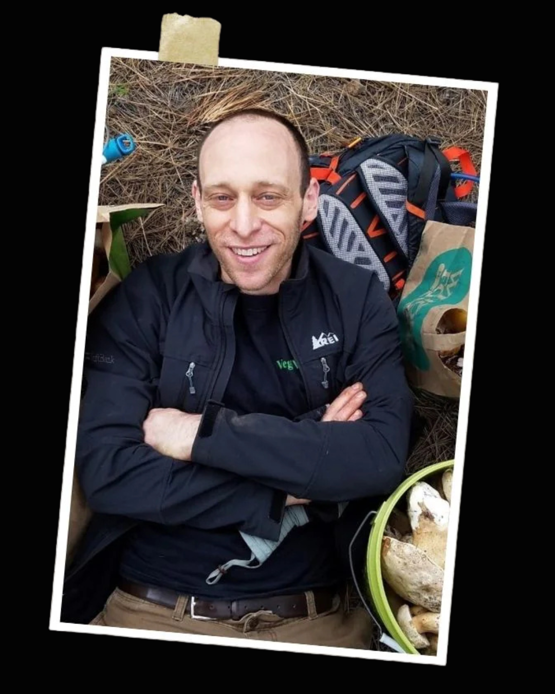 Man wearing a black jacket with a logo, sitting outdoors with a colorful blanket and plants.