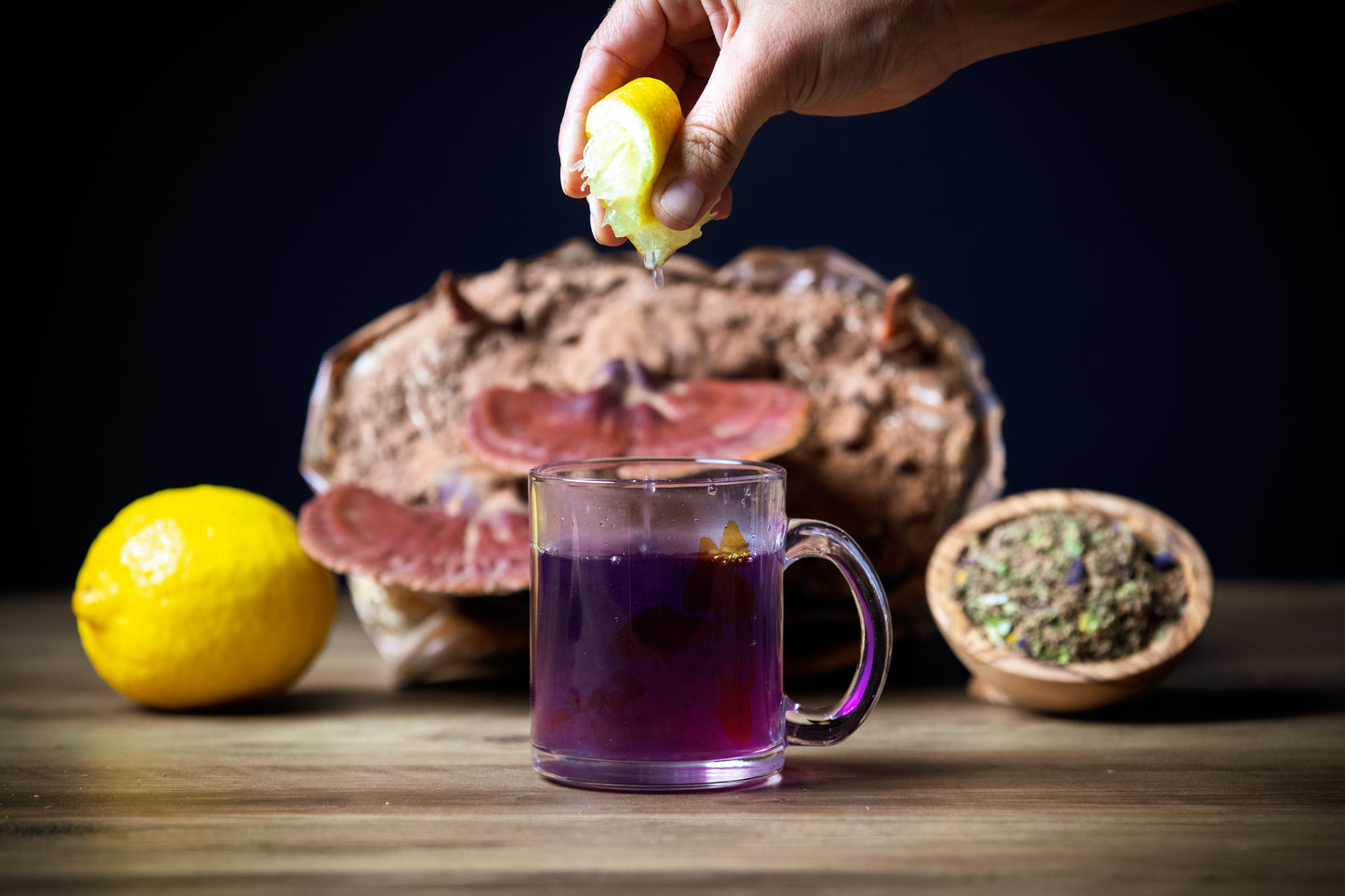 Hand squeezing lemon over a glass of purple liquid with a loaf of bread and lemons in the background.
