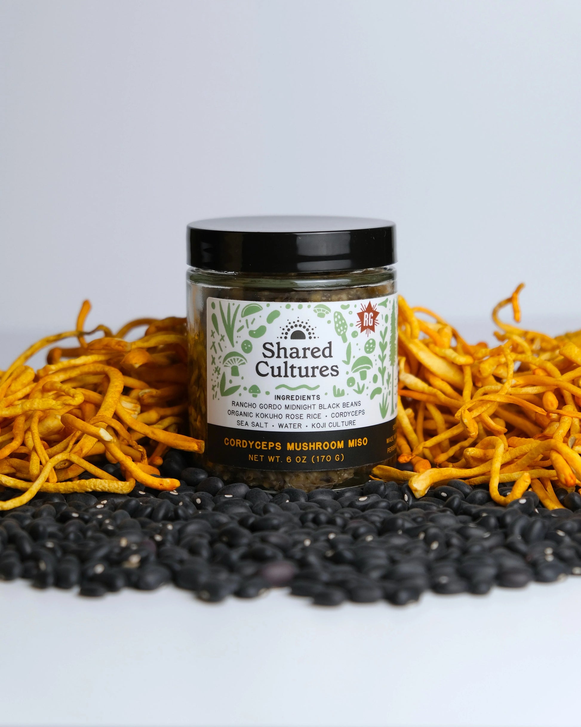 Jar of 'Shared Cultures' mushroom mix surrounded by various mushrooms on a light background